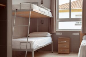 A clean and minimalistic hostel room with a bunk bed, white bedding, and small wooden bedside table